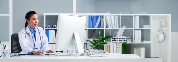 asian-doctor-with-stethoscope-around-neck-sitting-office-working-computer-EXT